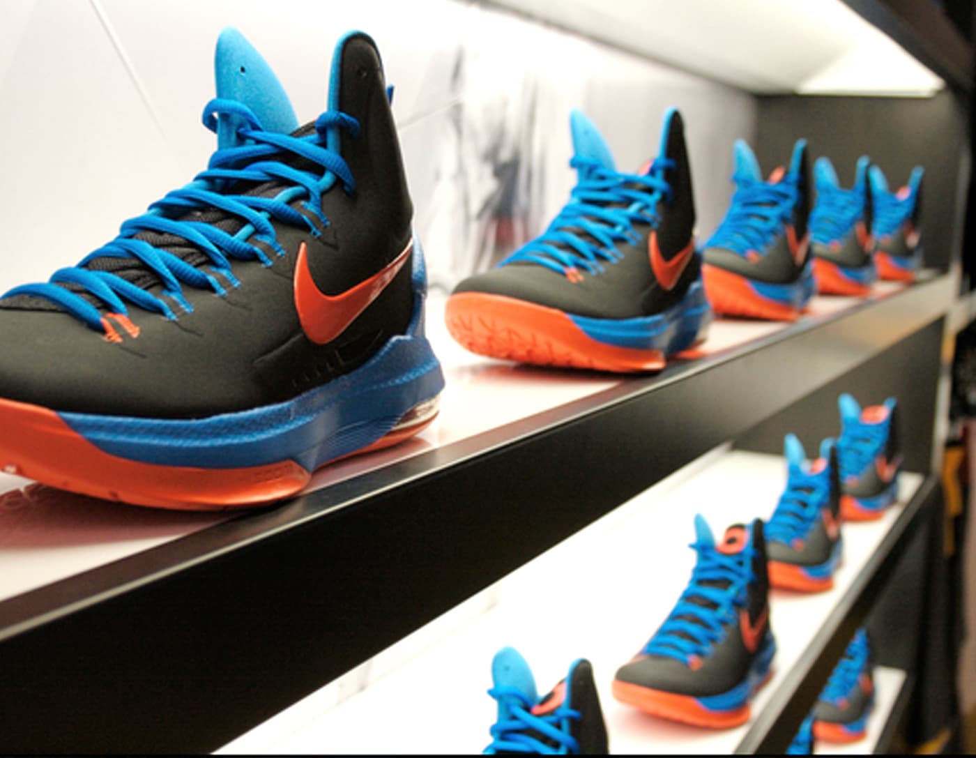 Rows of Kevin Durant Nike basketball shoes, shown in black with bright blue laces, orange Swoosh logos and orange soles, displayed on illuminated shelves inside a House of Hoops retail store.