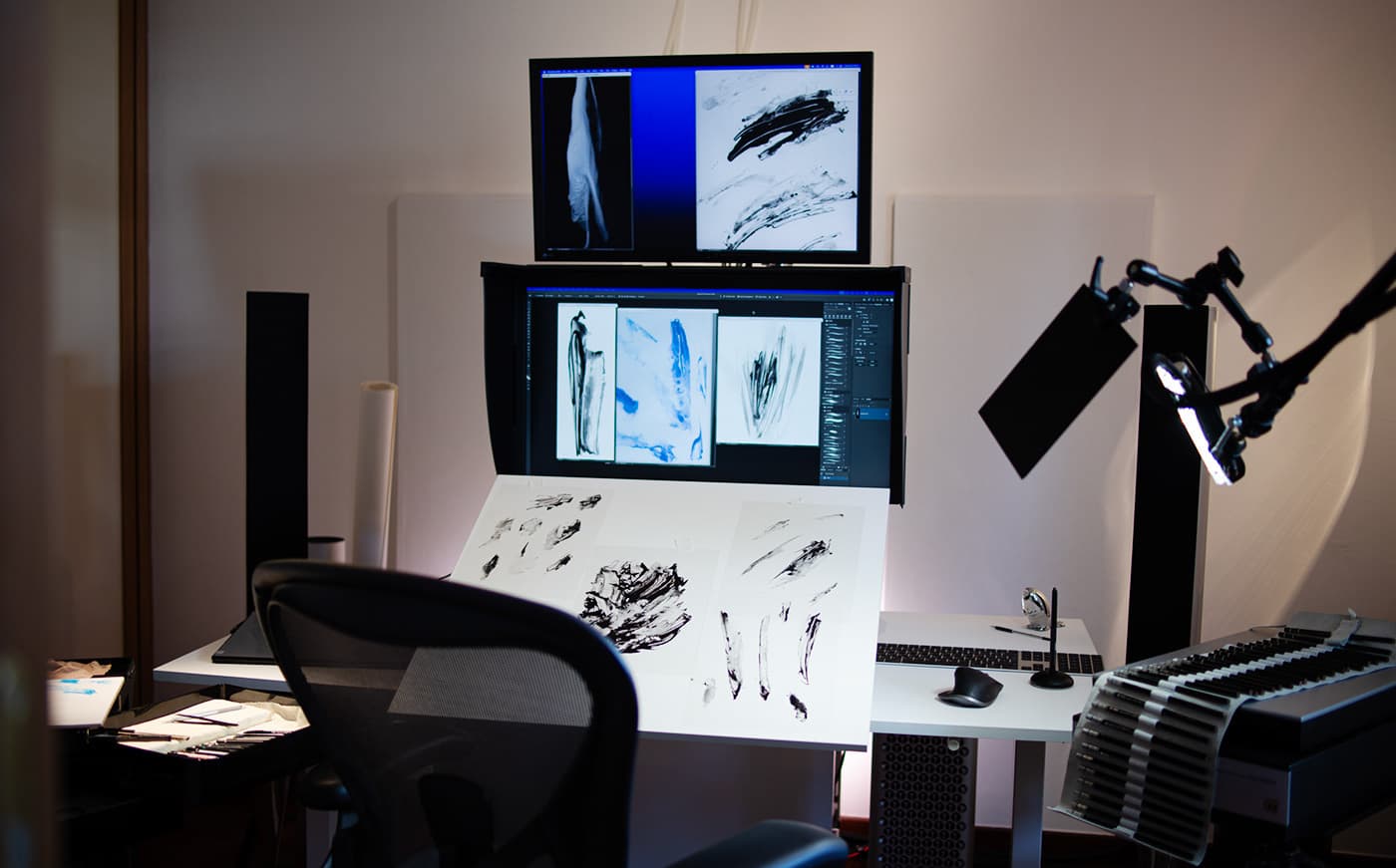 Alexis Marcou’s studio workspace, with painted studies on a tilted drawing board and two stacked screens displaying artwork for Atomic Maverick skis.