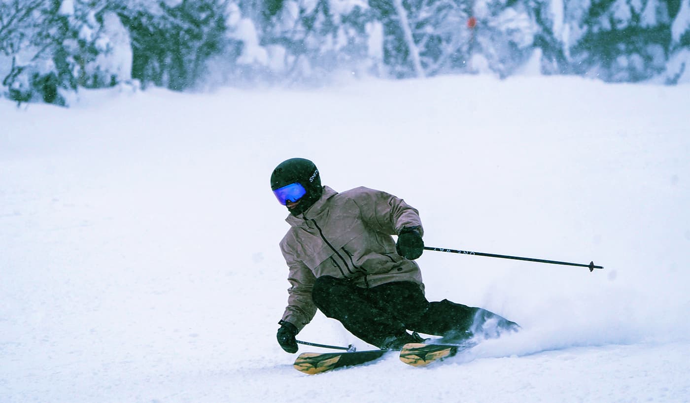 Skier carving through powder on yellow Atomic Maverick skis with black abstract graphics, spraying snow against a backdrop of snow-covered trees.