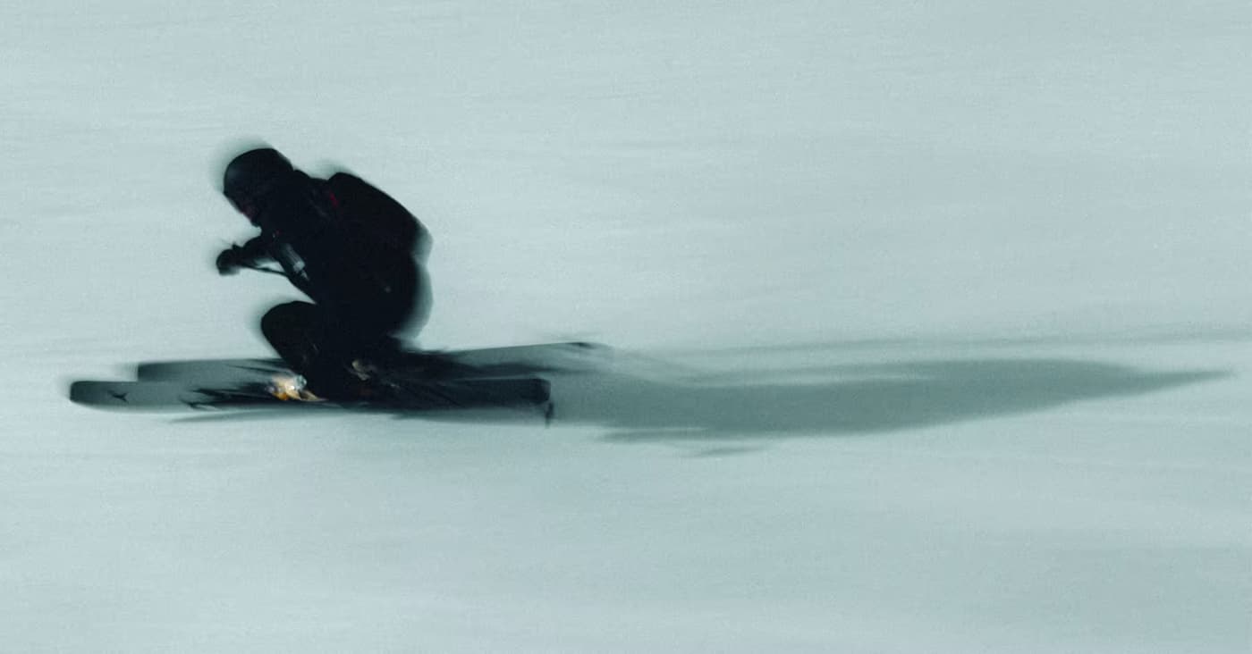 A skier in dark winter gear performs a jump on Atomic Maverick skis against a snowy mountain backdrop, captured with motion blur and a long shadow stretching across the snow.