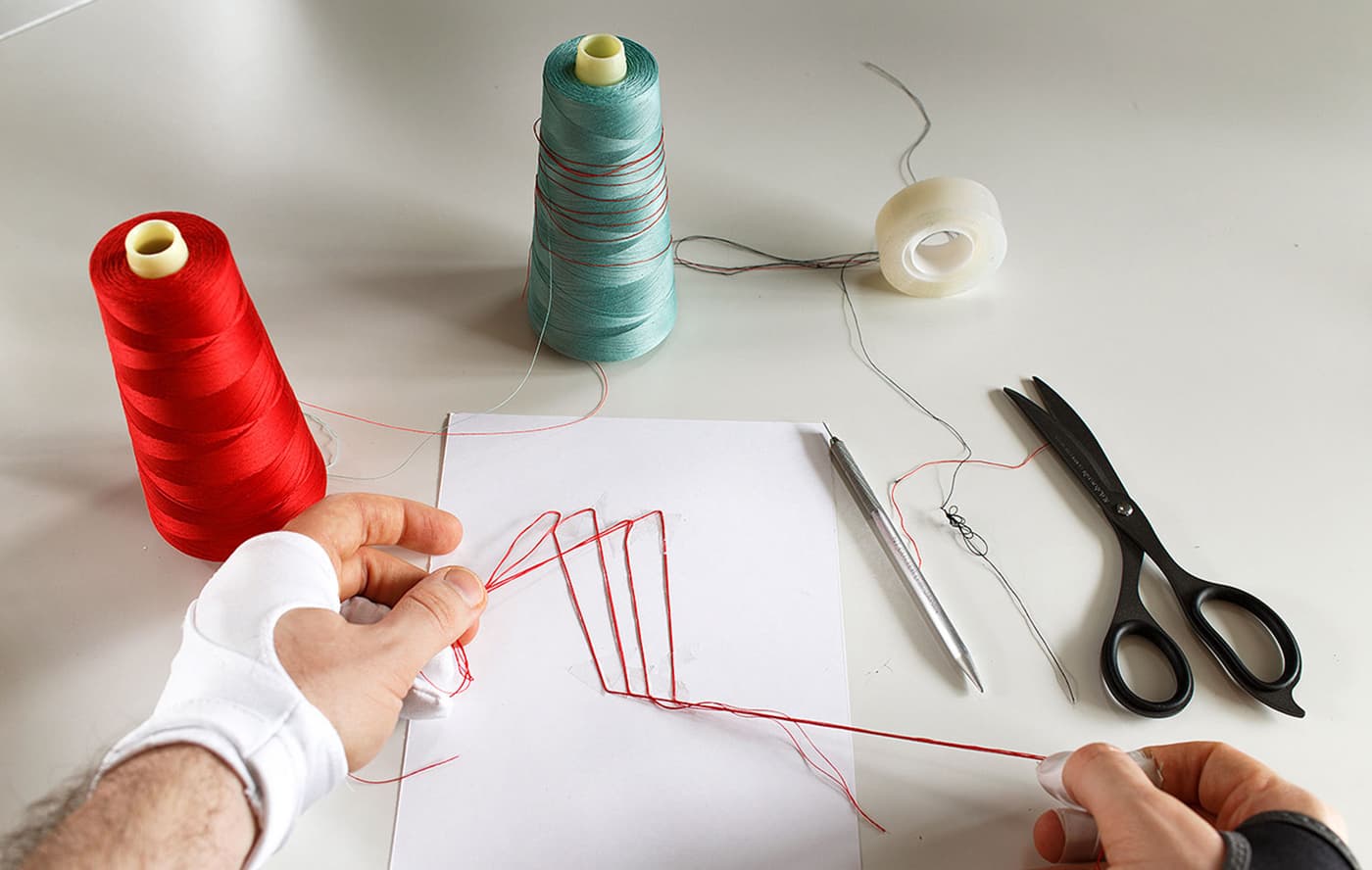 Hands shaping red thread around geometric line studies with thread spools, scissors and drawing tools on a studio table.
