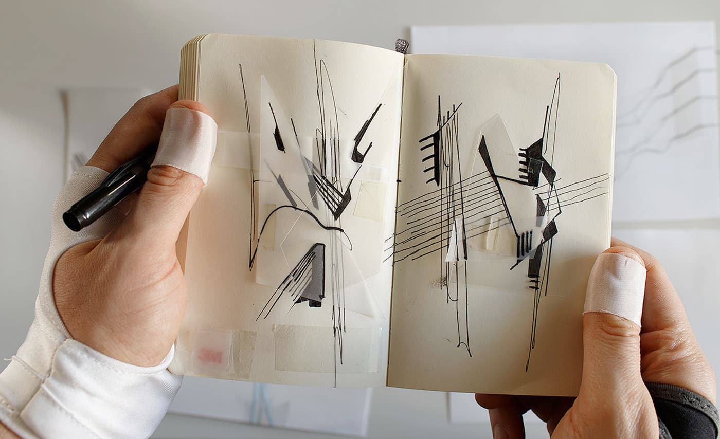 Hands holding an open sketchbook with black geometric line studies and translucent paper overlays.