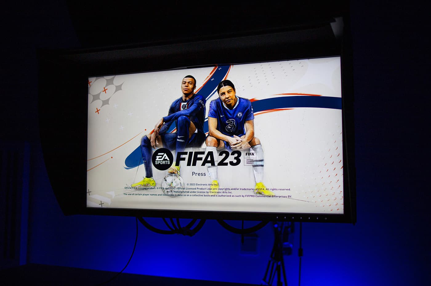 EA Sports FIFA 23 title screen showing the cover artwork with Kylian Mbappé and Sam Kerr on a monitor.