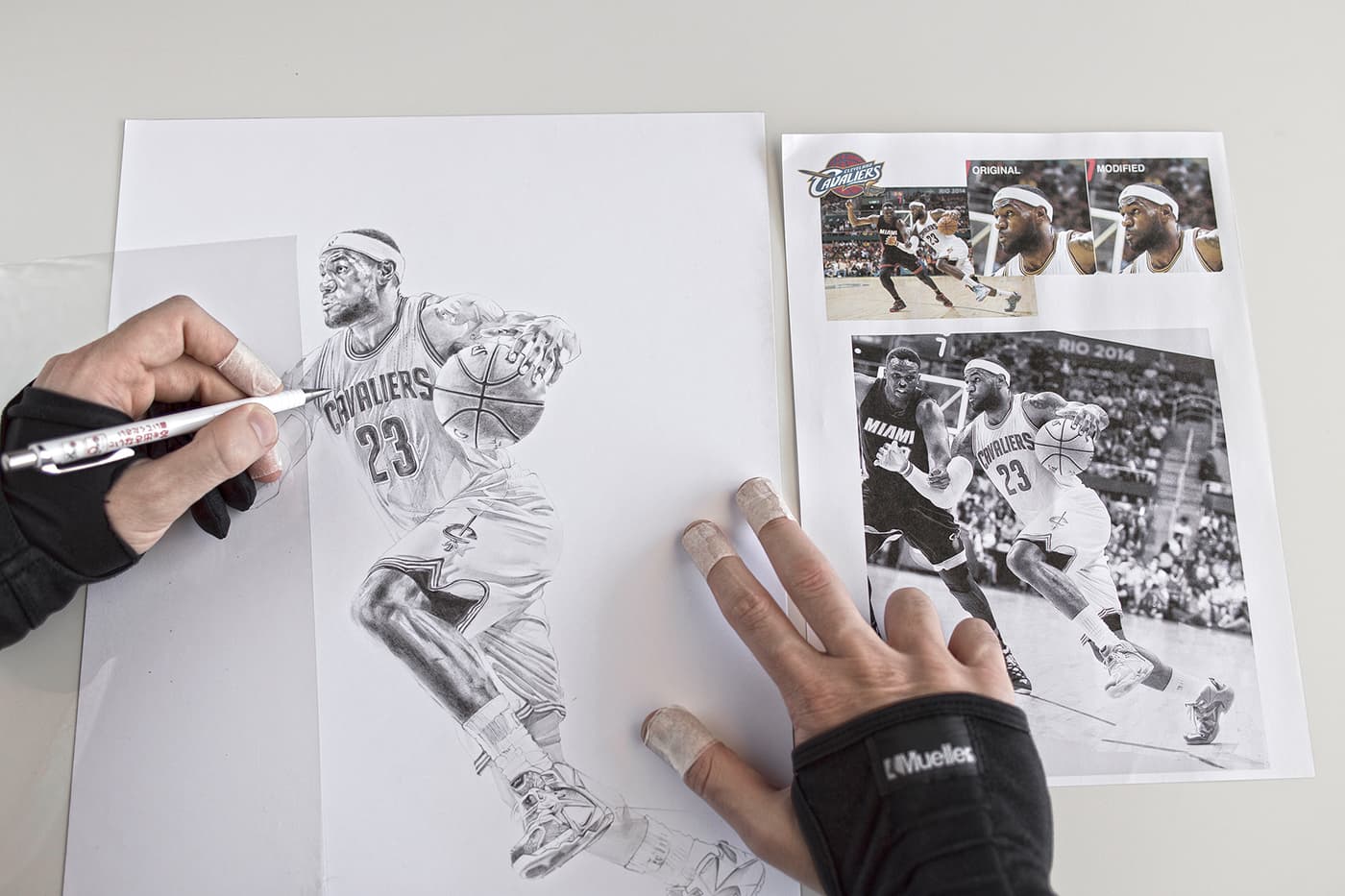 Hands working on a pencil illustration of LeBron James in a Cleveland Cavaliers uniform, with printed basketball reference photographs alongside the drawing.